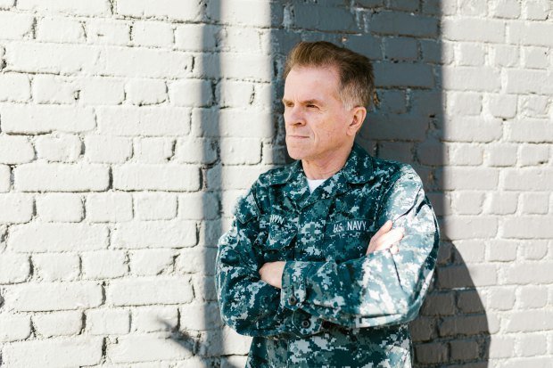 Senior US Navy veteran in uniform leaning against a sunlit brick wall.